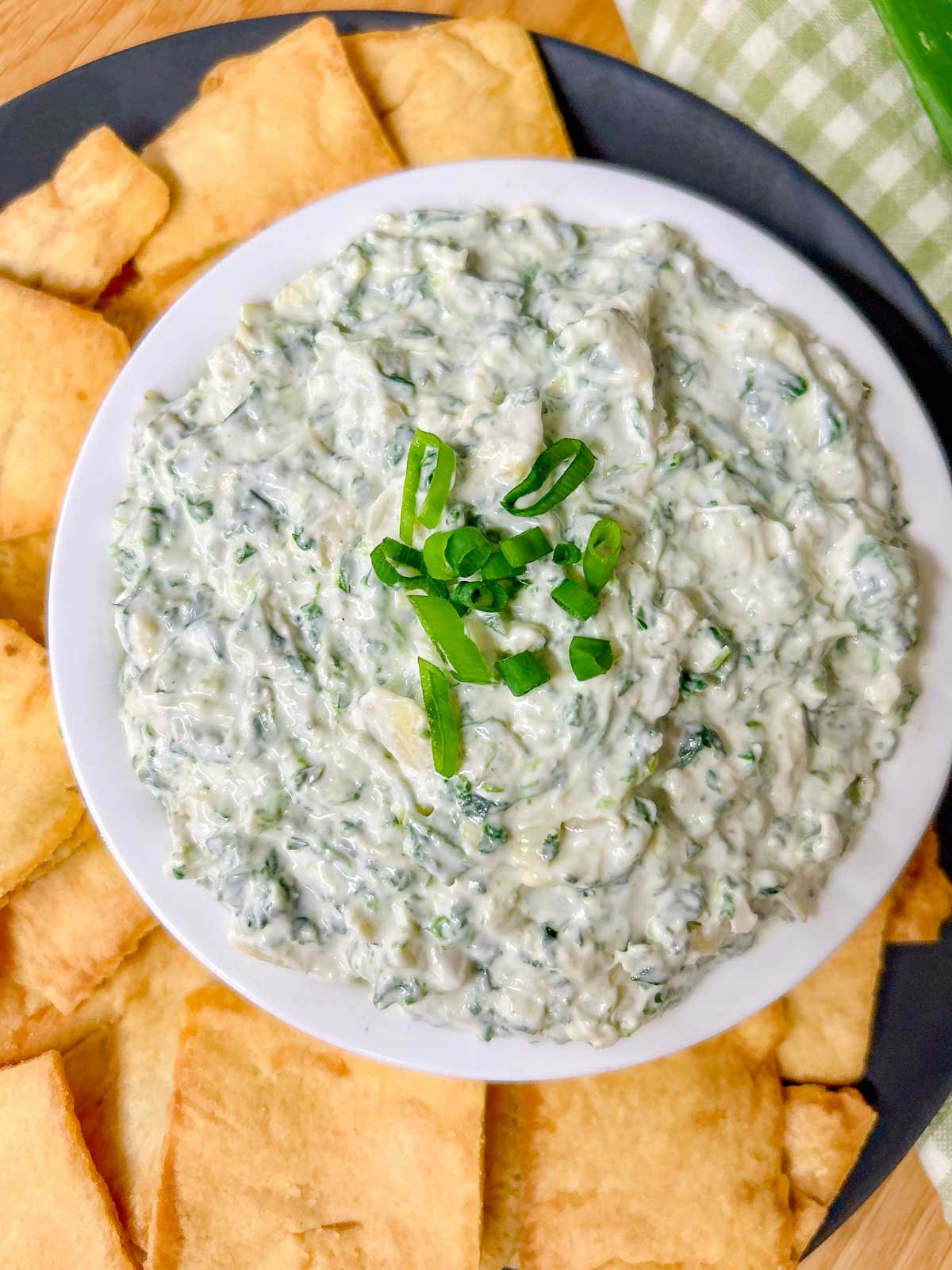 a black plate has a white serving bowl with spinach artichoke dip in it and pita chips around the bowl.