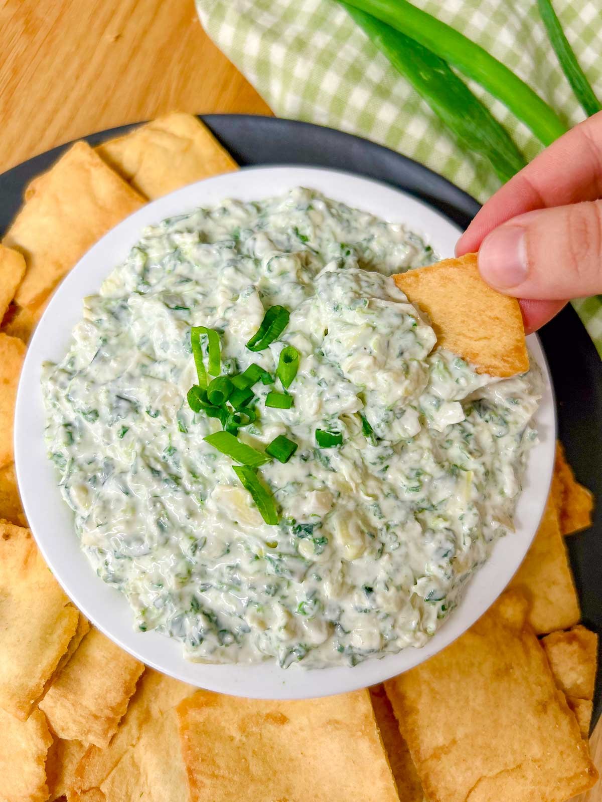a black plate has a white serving bowl with spinach artichoke dip in it and pita chips around the bowl. a hand is dipping a chip in the dip.