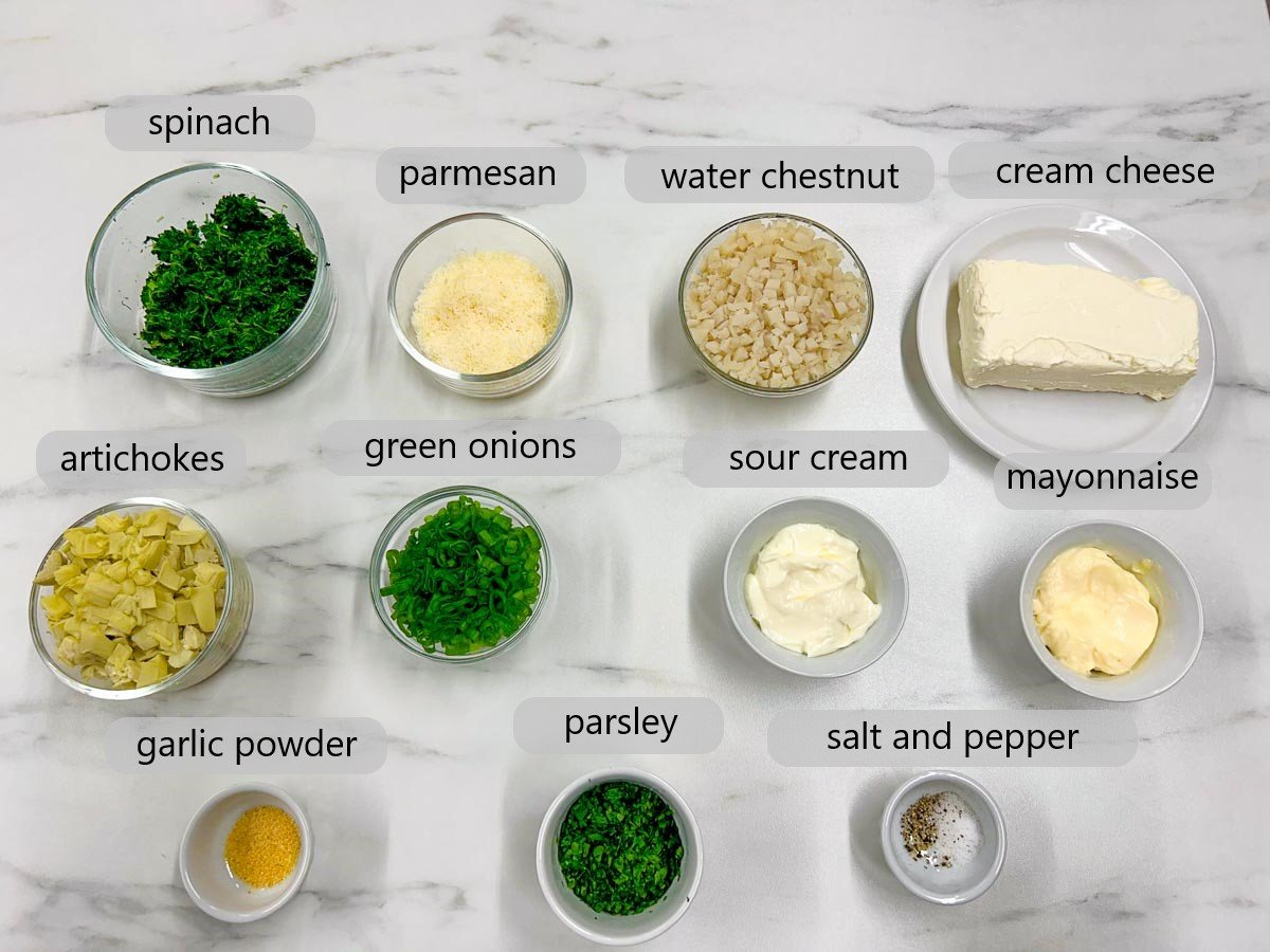 a white marble counter top has bowls with ingredients names labelled above them reading spinach, parmesan, water chestnuts, cream cheese, artichokes, green onions, sour cream, mayonnaise, artichokes, green onions, sour cream, mayonnaise, garlic powder, parsley, salt and pepper.