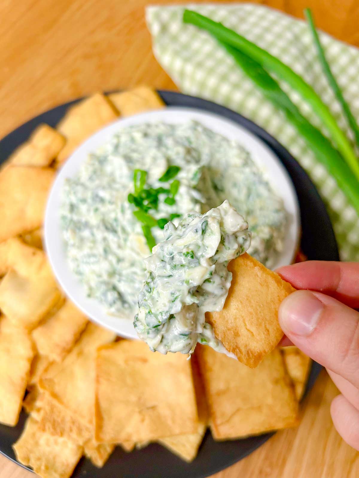 a black plate has a white serving bowl with spinach artichoke dip in it and pita chips around the bowl. one chip is dipped and held closer to the camera.