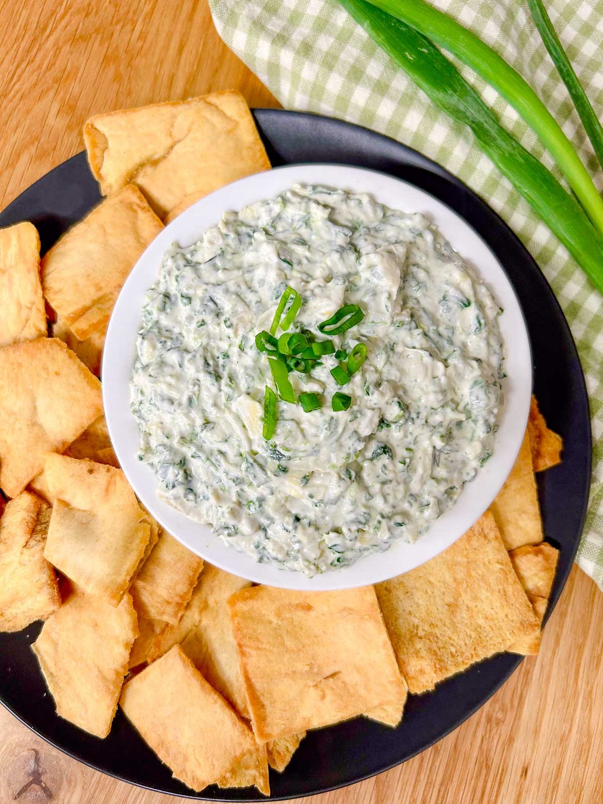 a black plate has a white serving bowl with spinach artichoke dip in it and pita chips around the bowl.