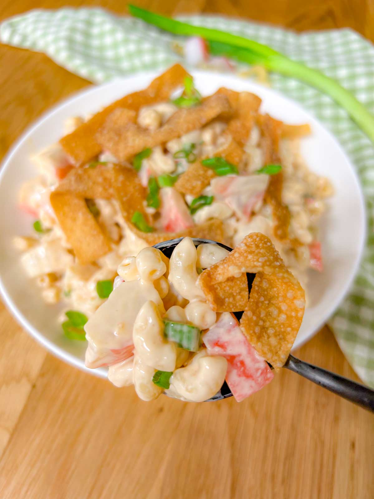 a white bowl is filled with crab rangoon pasta salad topped with crispy wonton strips and green onions. a spoon holds a spoonful of salad closer to the camera.