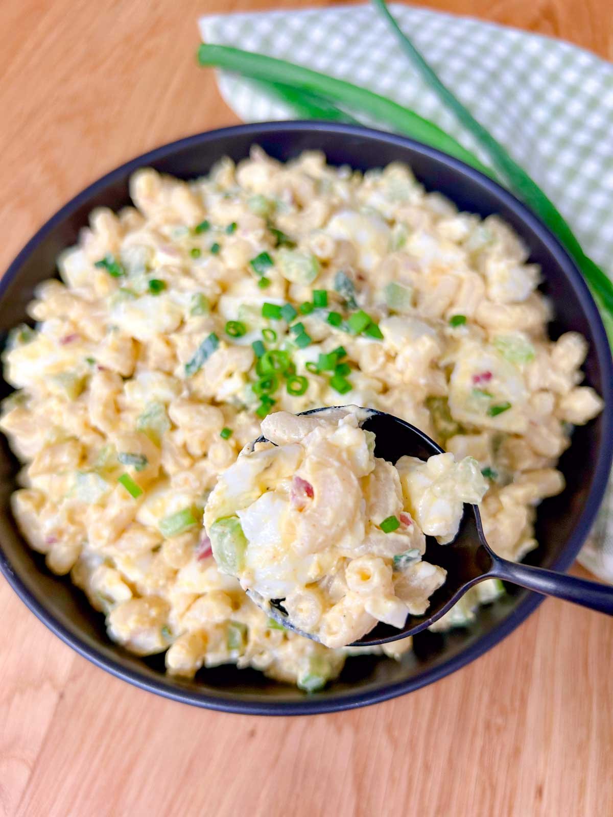 a black bowl is filled with deviled egg pasta salad and garnished with green onions. a spoon is held close to the camera.