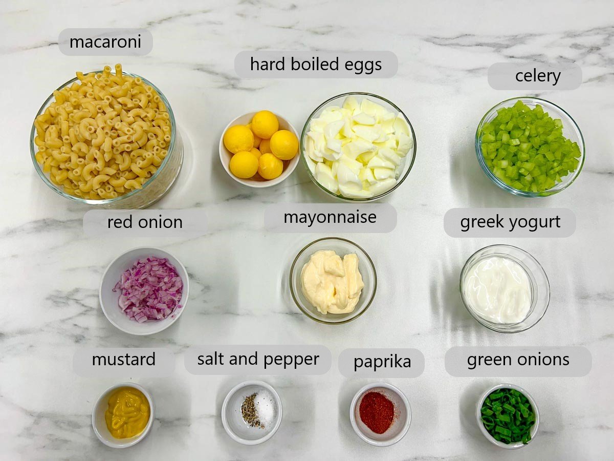 a white marble counter top has bowls with ingredients and text overlays reading macaroni, hard boiled eggs, celery, red onion, mayonnaise, Greek yogurt, mustard, salt and pepper, paprika, and green onions.