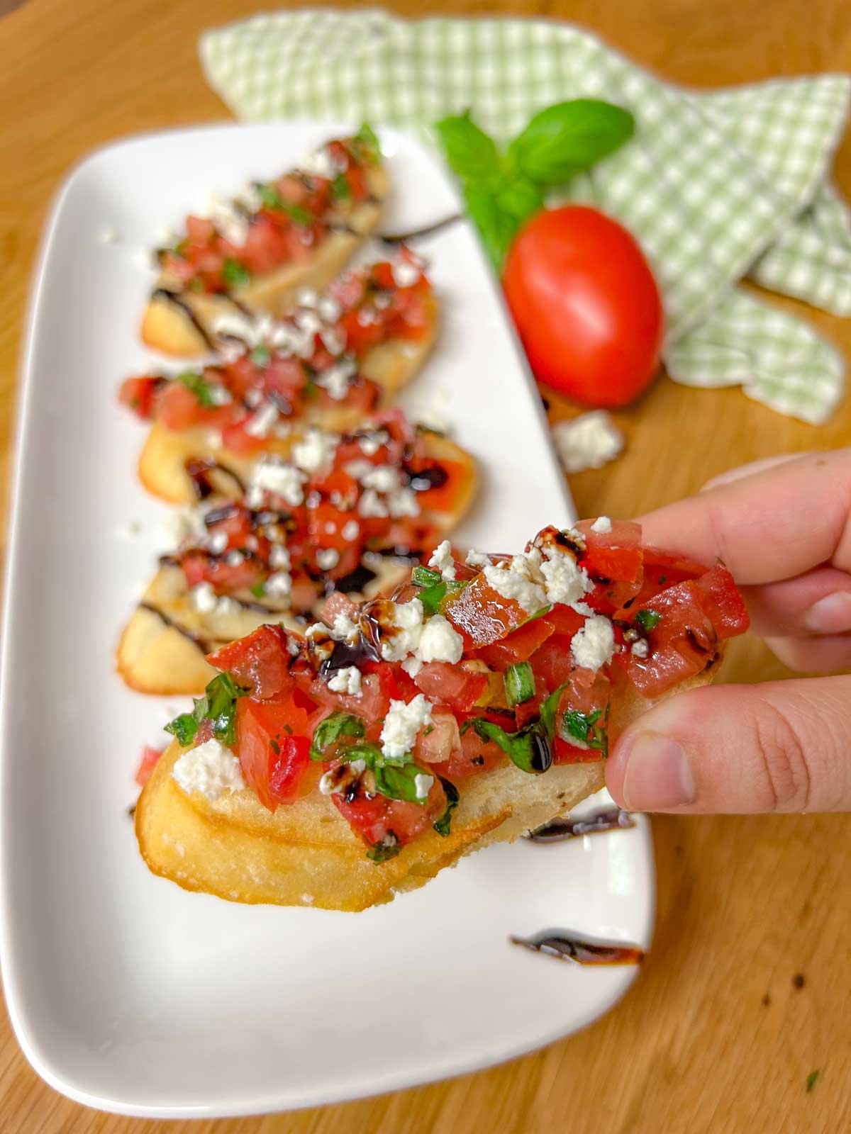 a white serving platter has 4 pieces of bruschetta with crumbled feta cheese and balsamic glaze on top. one piece is held up close to the camera.