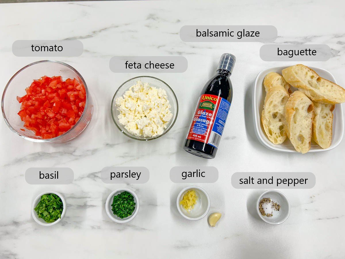 a white countertop has bowls with ingredients in them with text labels reading tomatoes, feta cheese, balsamic glaze, baguette, basil, parsley, garlic, salt, and pepper.