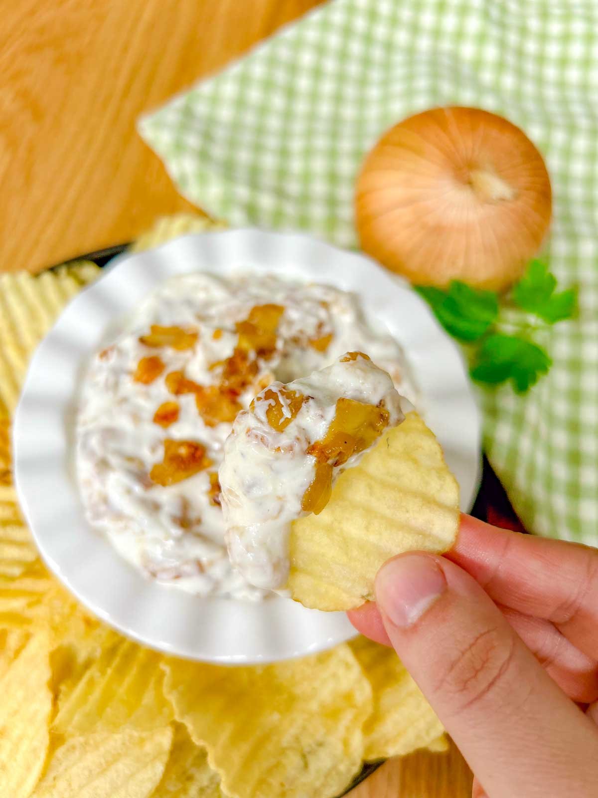 a white bowl is filled with French onion dip and garnished with caramelized onions. a chip is dipped in the dip and held up close to the camera.