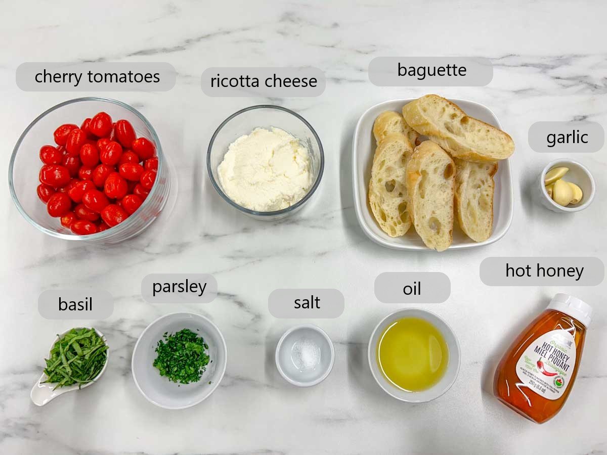 a white marble kitchen counter top has bowls with the ingredients to make hot honey roasted bruschetta with the ingredients labelled as cherry tomaotes, ricotta cheese, baguette, garlic, basil, parsley, salt, oil and hot honey.
