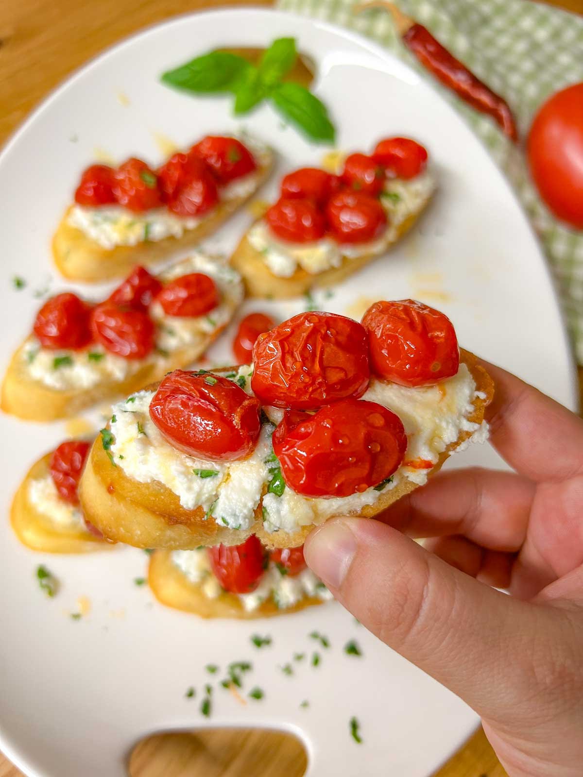 a white serving platter has pieces of hot honey roasted bruschetta on top and is garnished with hot honey, basil, and parsley. one piece of bruschetta is being held up close to the camera.