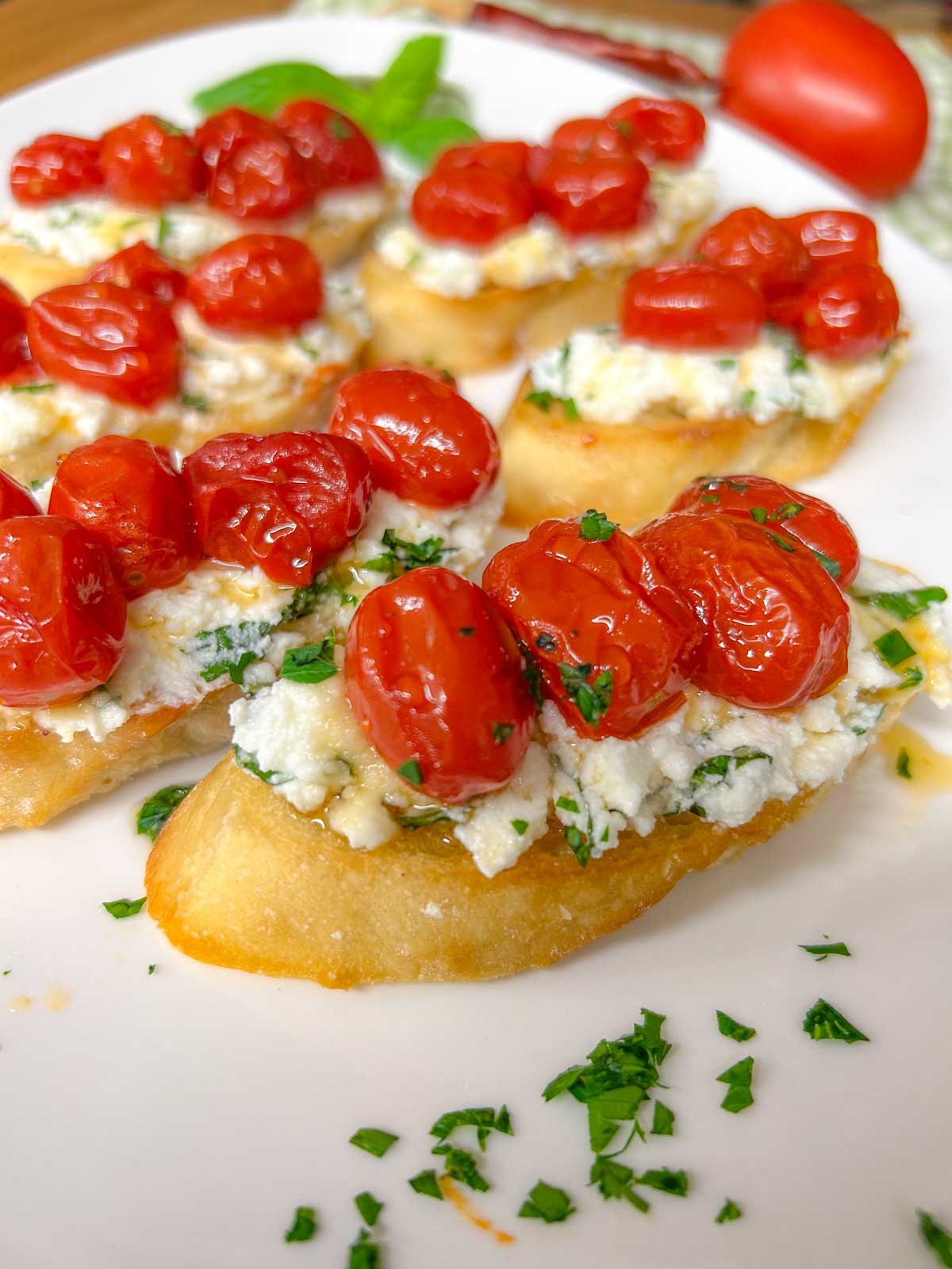 a white serving platter has pieces of hot honey roasted bruschetta on top and is garnished with hot honey, basil, and parsley.