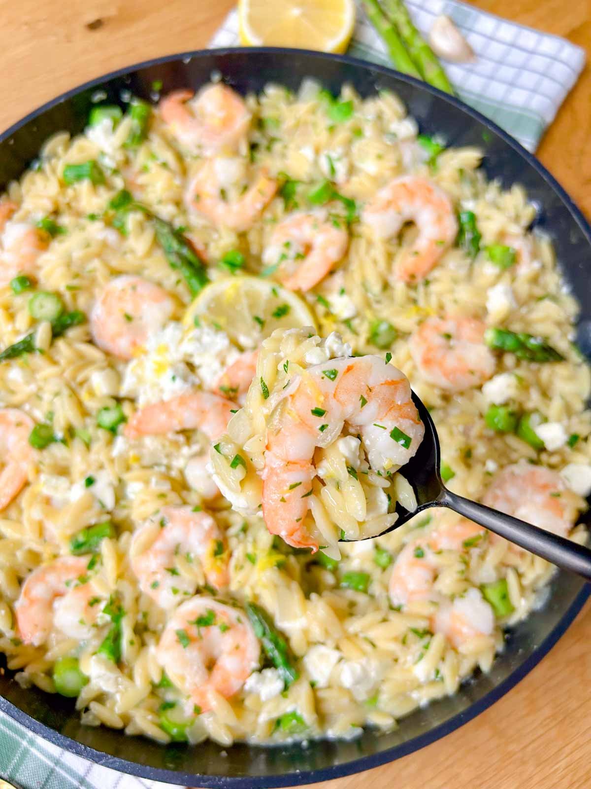 a black frying pan is filled with lemon feta shrimp orzo and garnished with parsley. a black spoon is holding a spoonful of the orzo pasta and a shrimp.