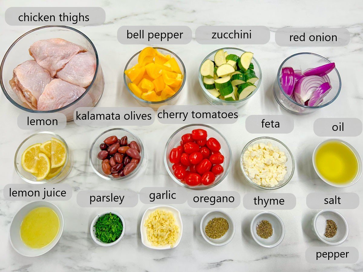 a white marble counter top has bowls with the ingredients to make Greek sheet pan dinner with text overlay reading chicken thighs, bell pepper, zucchini, red onion, lemon, kalamata olives, cherry tomatoes, feta, oil, lemon juice, parsley, garlic, oregano, thyme, salt and pepper.