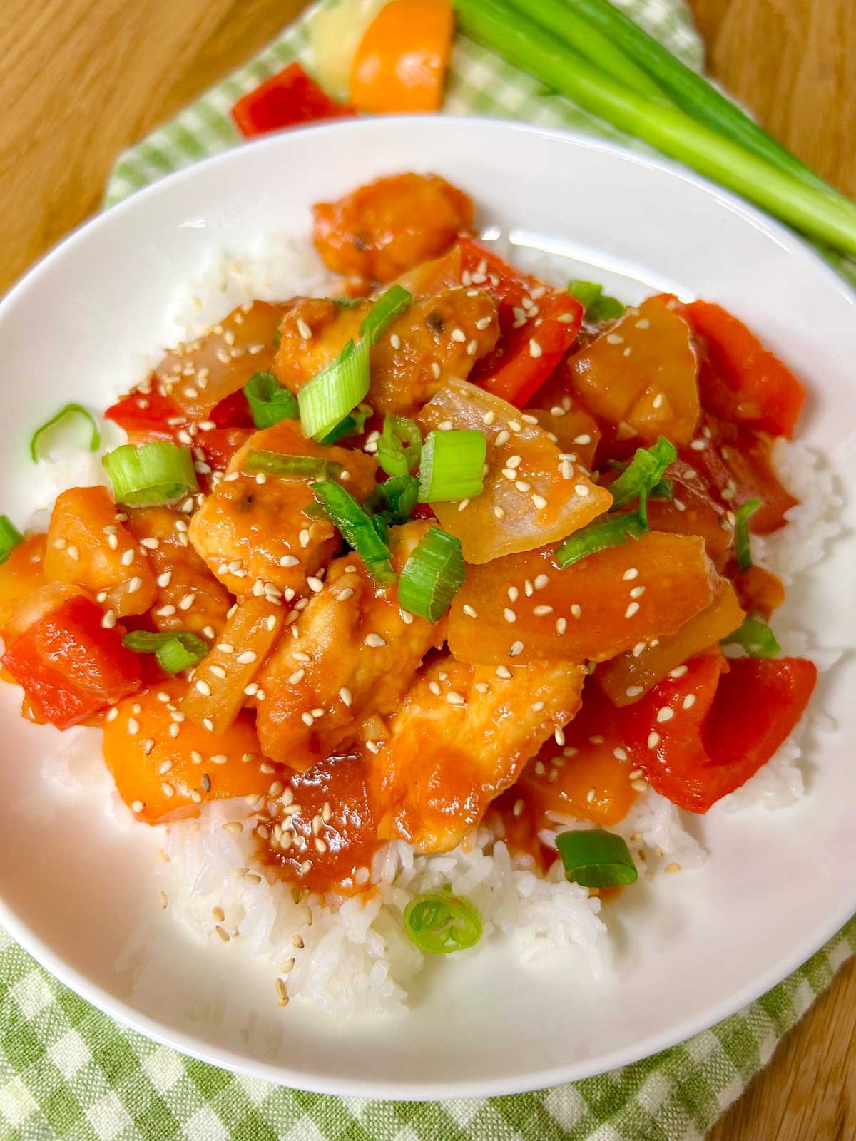 a white bowl is filled with white rice and topped with a sweet and sour chicken stir fry. it is garnished with sesame seeds and sliced green onions