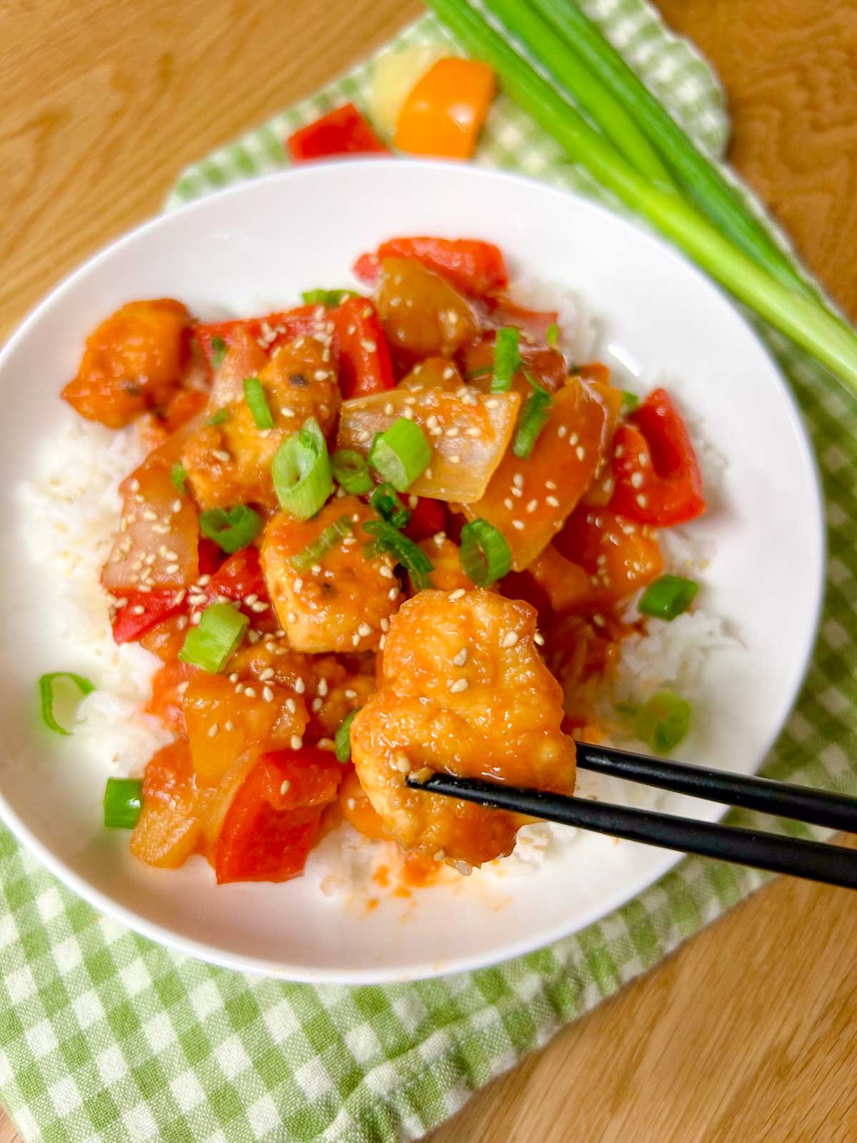 a white bowl is filled with white rice and topped with a sweet and sour chicken stir fry. it is garnished with sesame seeds and sliced green onions. black chopsticks are holding up a piece of chicken