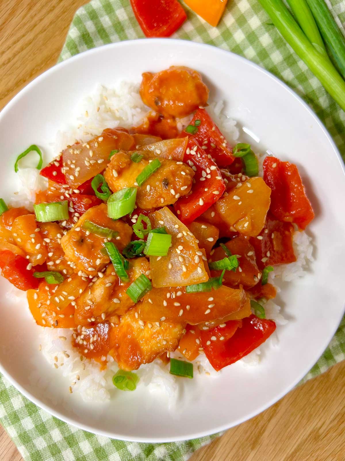 a white bowl is filled with white rice and topped with a sweet and sour chicken stir fry. it is garnished with sesame seeds and sliced green onions
