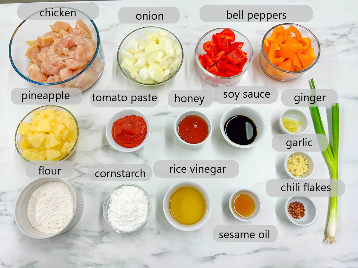 a white marble countertop has bowls with the ingredients to make sweet and sour chicken labelled - chicken, onion, bell peppers, pineapple, tomato paste, honey, soy sauce, ginger, garlic, flour, cornstarch, rice vinegar, sesame oil, and chili flakes are labelled with text overlay.