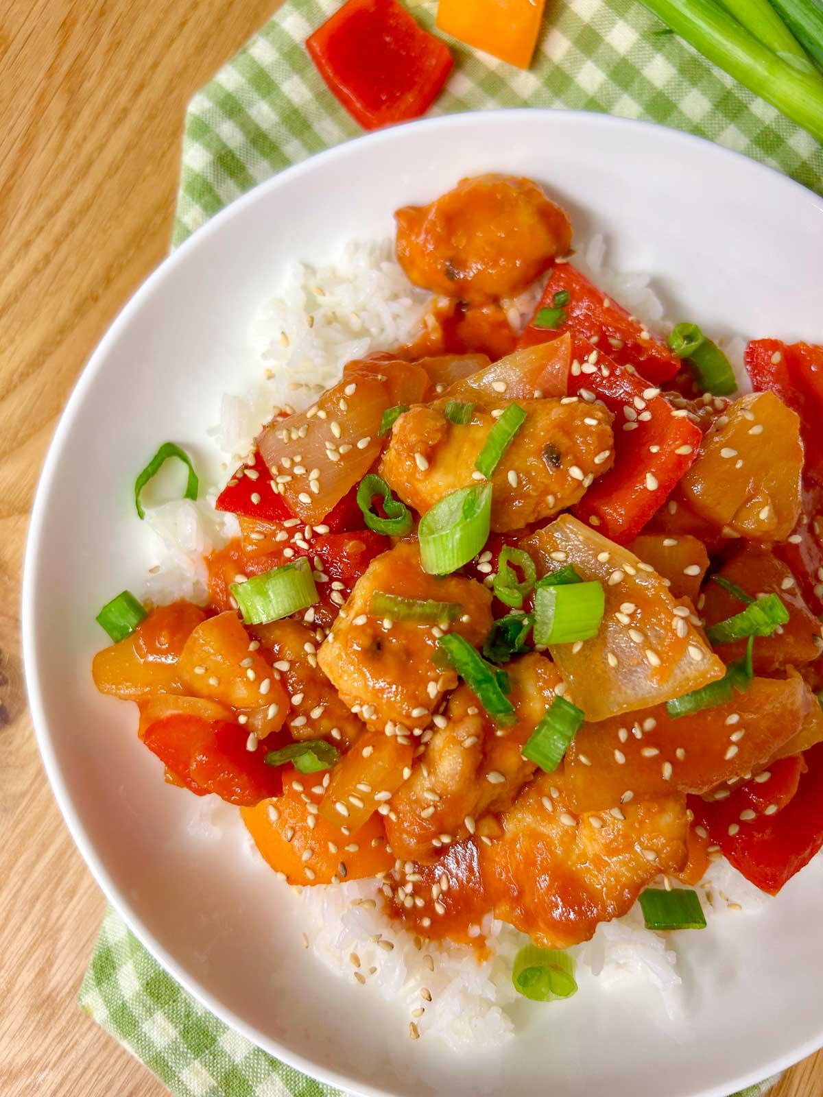 a white bowl is filled with white rice and topped with a sweet and sour chicken stir fry. it is garnished with sesame seeds and sliced green onions