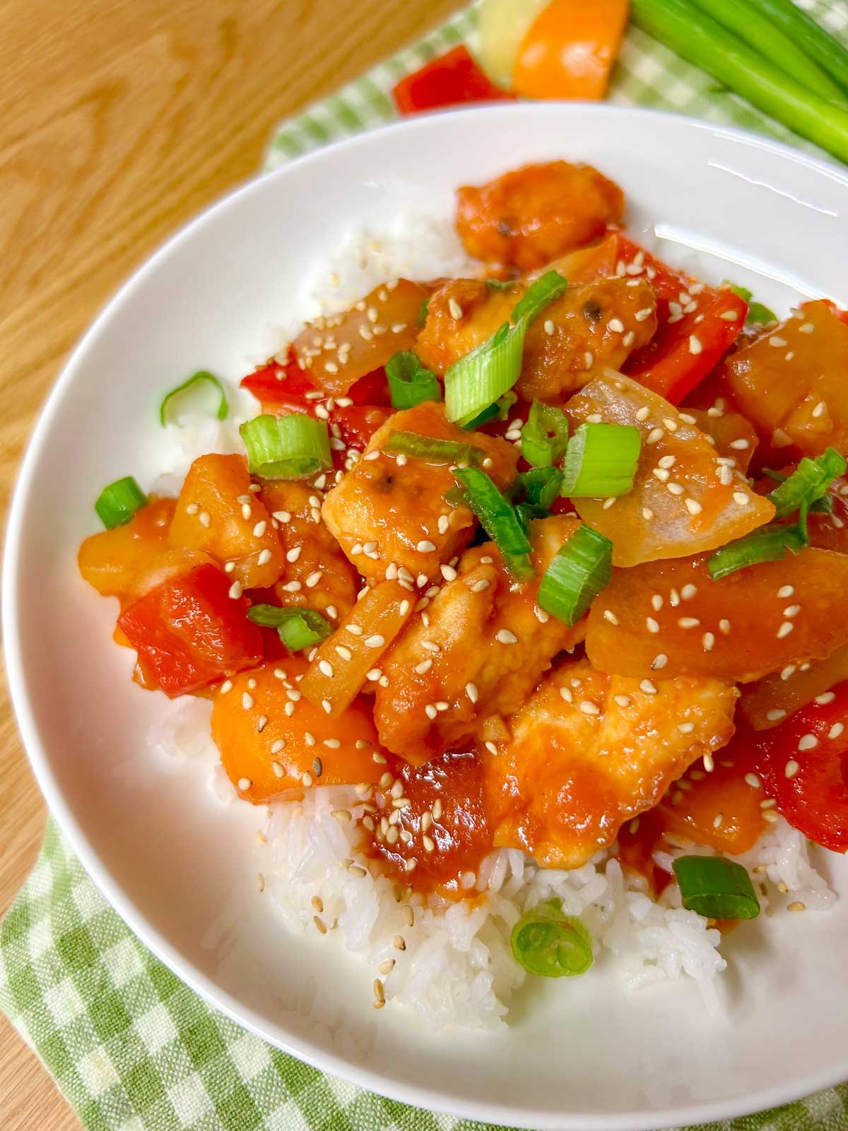 a white bowl is filled with white rice and topped with a sweet and sour chicken stir fry. it is garnished with sesame seeds and sliced green onions