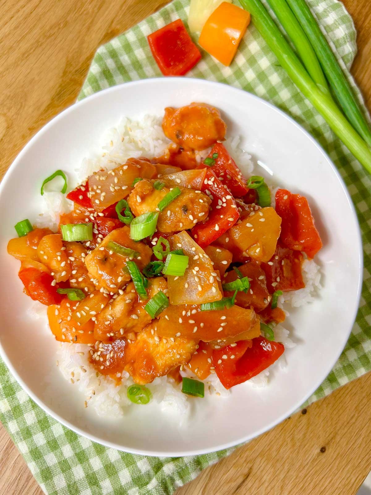 a white bowl is filled with white rice and topped with a sweet and sour chicken stir fry. it is garnished with sesame seeds and sliced green onions