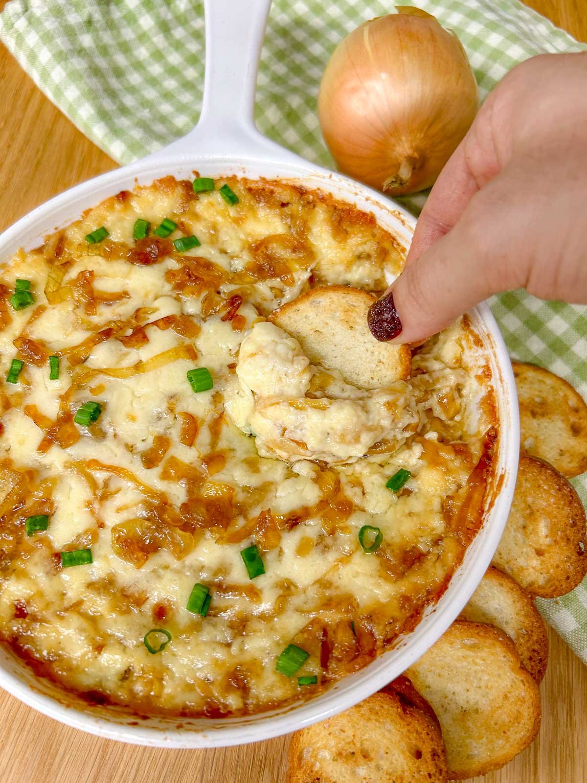 a white dish is filled with baked caramelized onion dip and garnished with green onions. a piece of bread is dipped