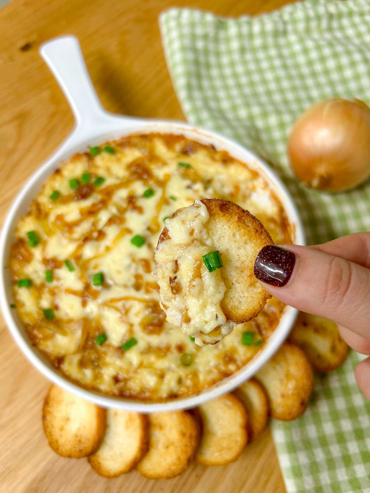 a white dish is filled with baked caramelized onion dip and garnished with green onions. a piece of dipped bread is held close to the camera