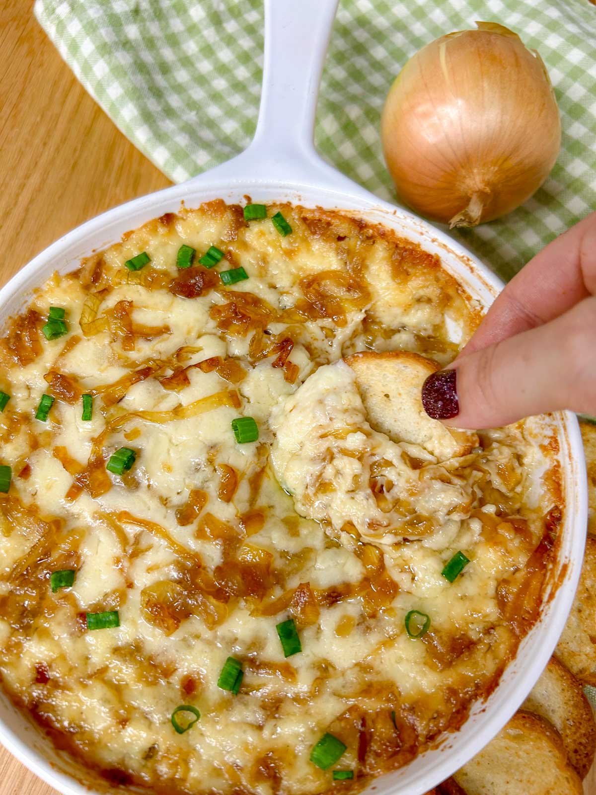 a white dish is filled with baked caramelized onion dip and garnished with green onions. a piece of bread is dipped in the dip