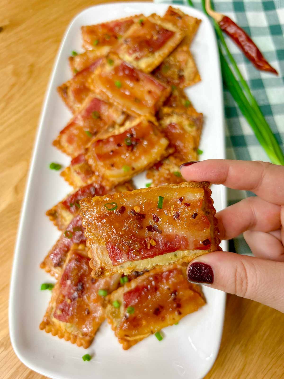 a white plate has candied bacon boursin crackers on top and garnished with green onions. one is held up close to the camera