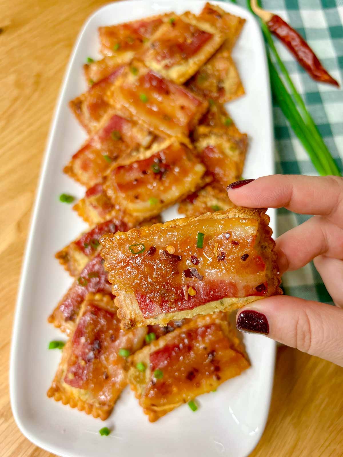 a white plate has candied bacon boursin crackers on top and garnished with green onions. one is held close to the camera