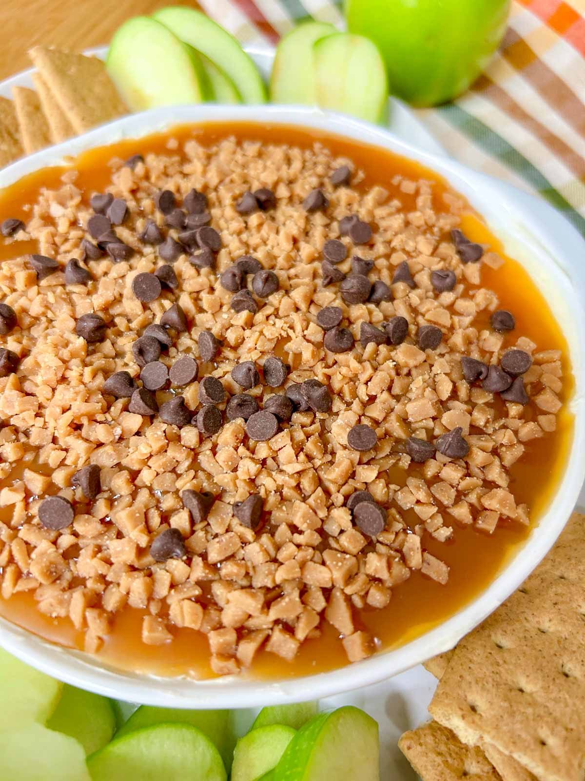 a white bowl is filled with caramel apple cheesecake dip and served with apple and graham crackers to dip