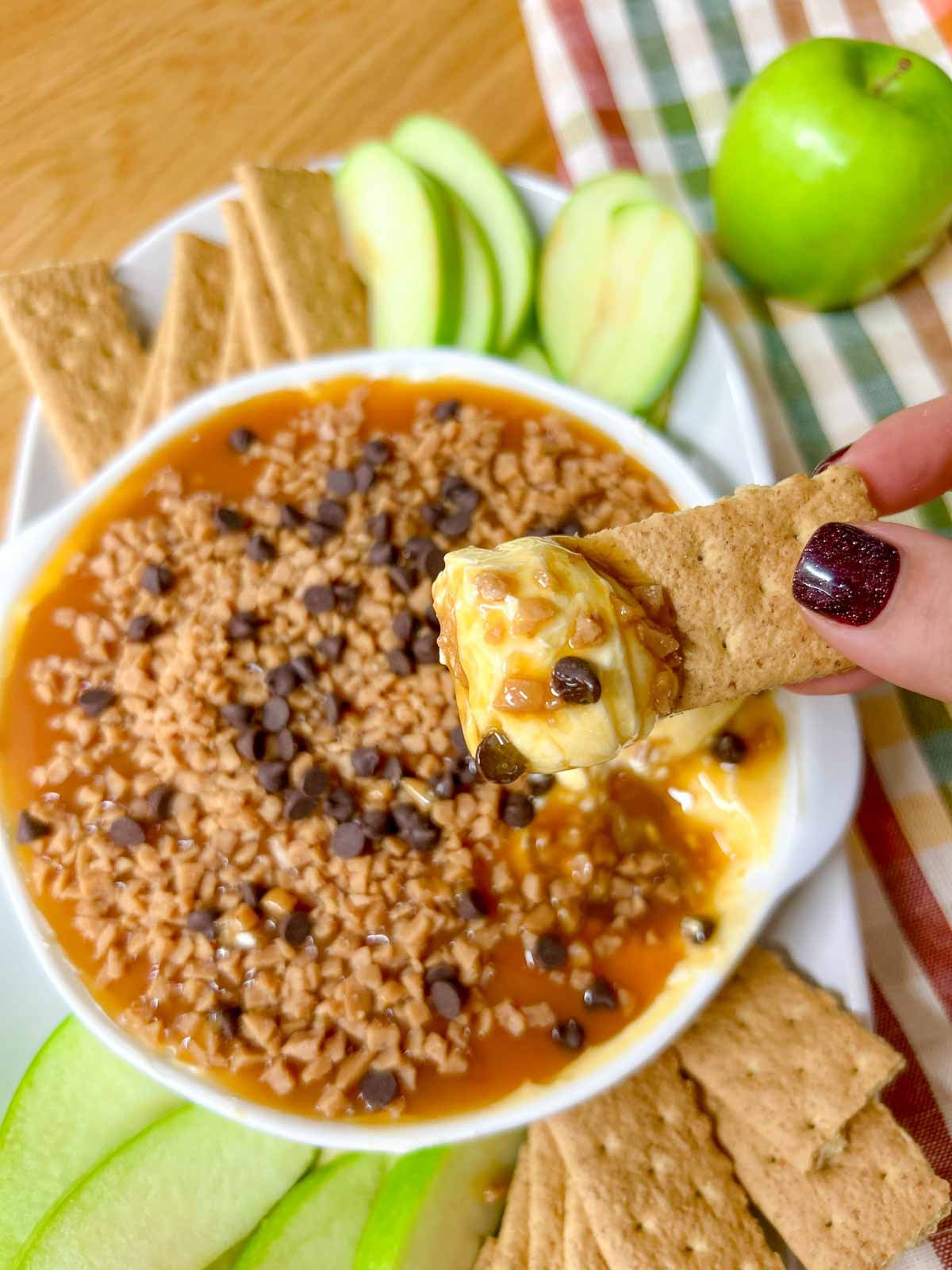 a white bowl is filled with caramel apple cheesecake dip and served with apple and graham crackers to dip. one graham cracker is dipped into the dip and held close to the camera