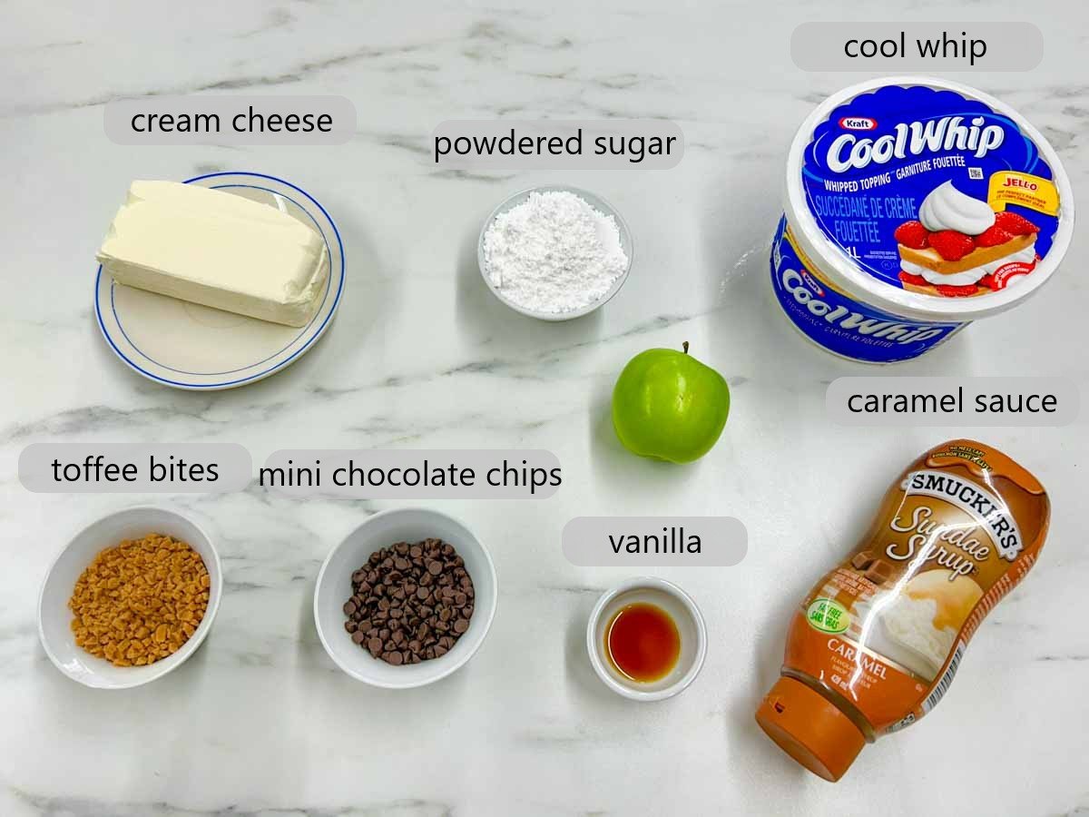 a kitchen counter with bowls of the ingredients to make caramel apple cheesecake dip and text overlays reading cream cheese, powdered sugar, cool whip, toffee bits, mini chocolate chips, vanilla, and caramel sauce