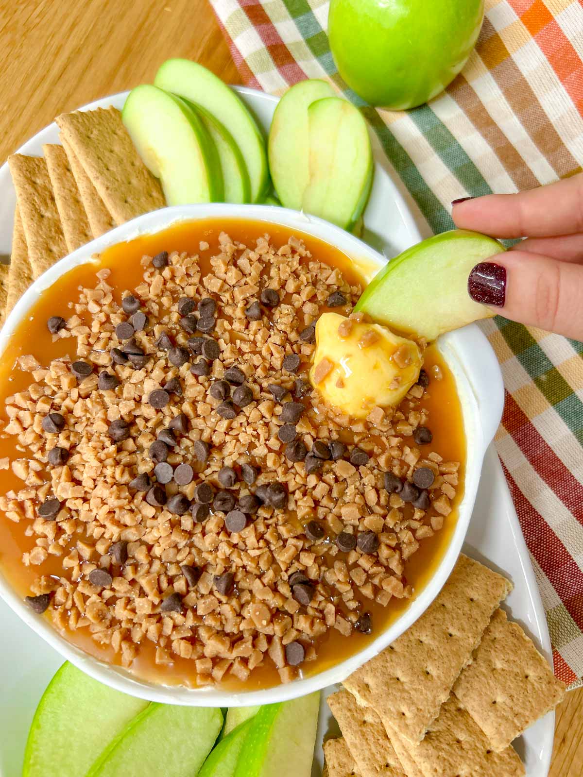 a white bowl is filled with caramel apple cheesecake dip and served with apple and graham crackers to dip. one apple is dipped into the dip
