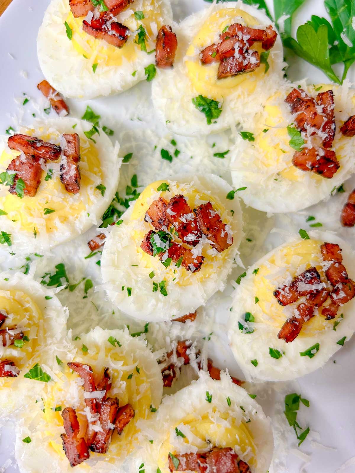 a white serving platter is filled with carbonara deviled eggs that are topped with crispy pancetta, grated parmesan cheese, and fresh parsley