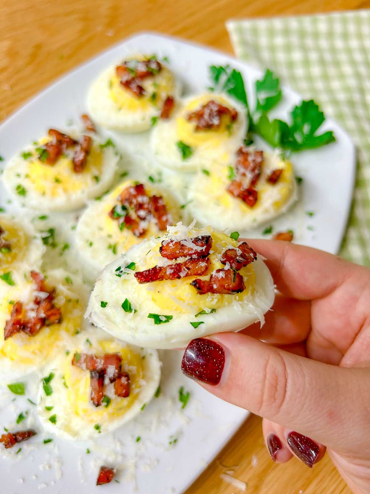 a white serving platter is filled with carbonara deviled eggs that are topped with crispy pancetta, grated parmesan cheese, and fresh parsley. one egg is held up to the camera