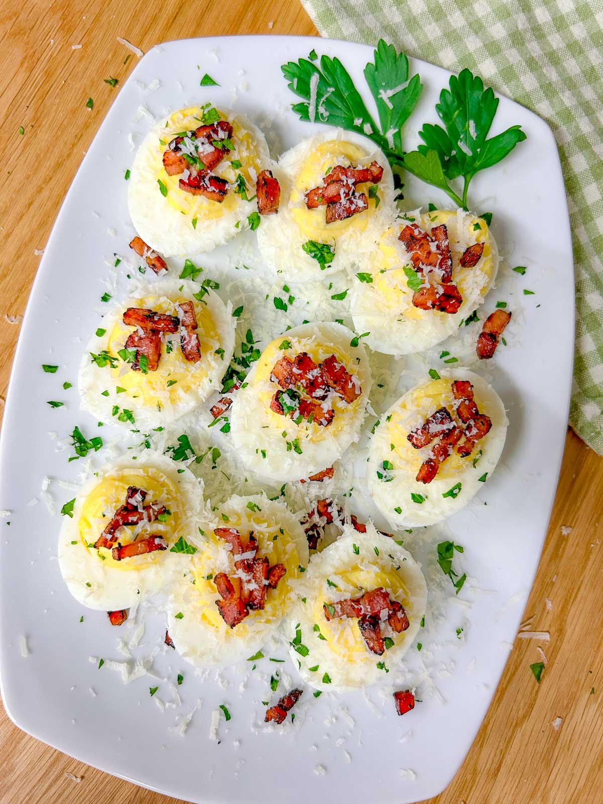 a white serving platter is filled with carbonara deviled eggs that are topped with crispy pancetta, grated parmesan cheese, and fresh parsley