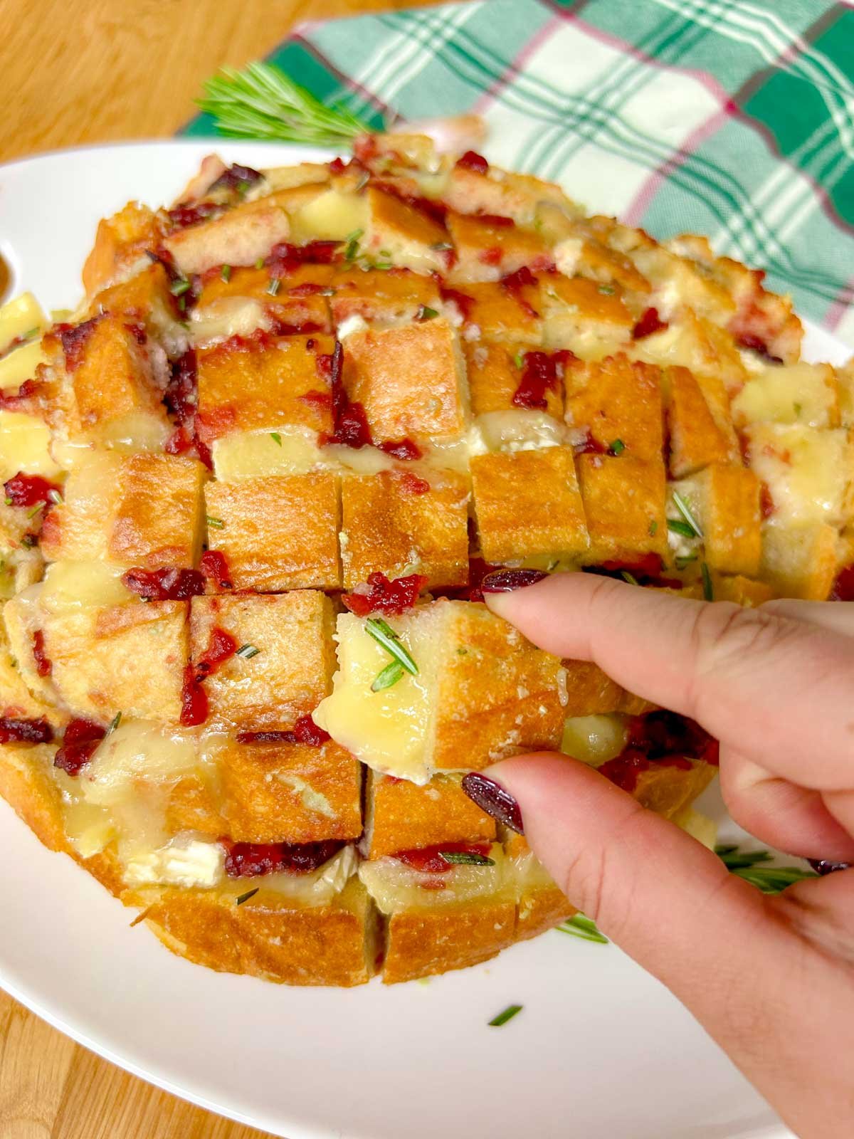 a loaf of cranberry brie pull apart garlic bread it on a white platter and garnished with rosemary. one piece is being pulled out