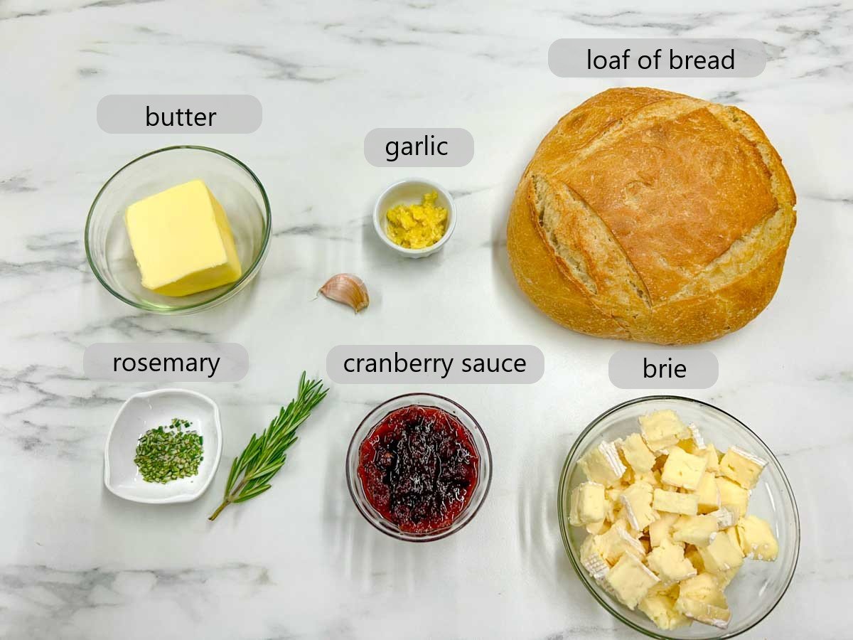 a kitchen counter with bowls of the ingredients to make cranberry brie pull apart garlic bread with text over lays reading brie, garlic, loaf of bread, rosemary, cranberry sauce and brie.