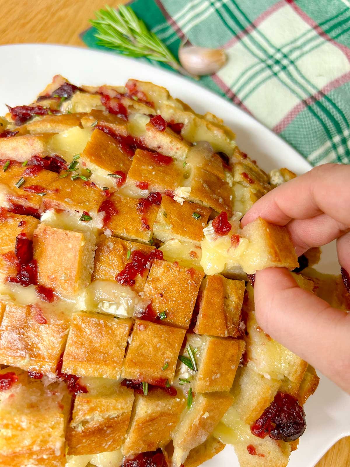 a loaf of cranberry brie pull apart garlic bread it on a white platter and garnished with rosemary. one piece is pulled out