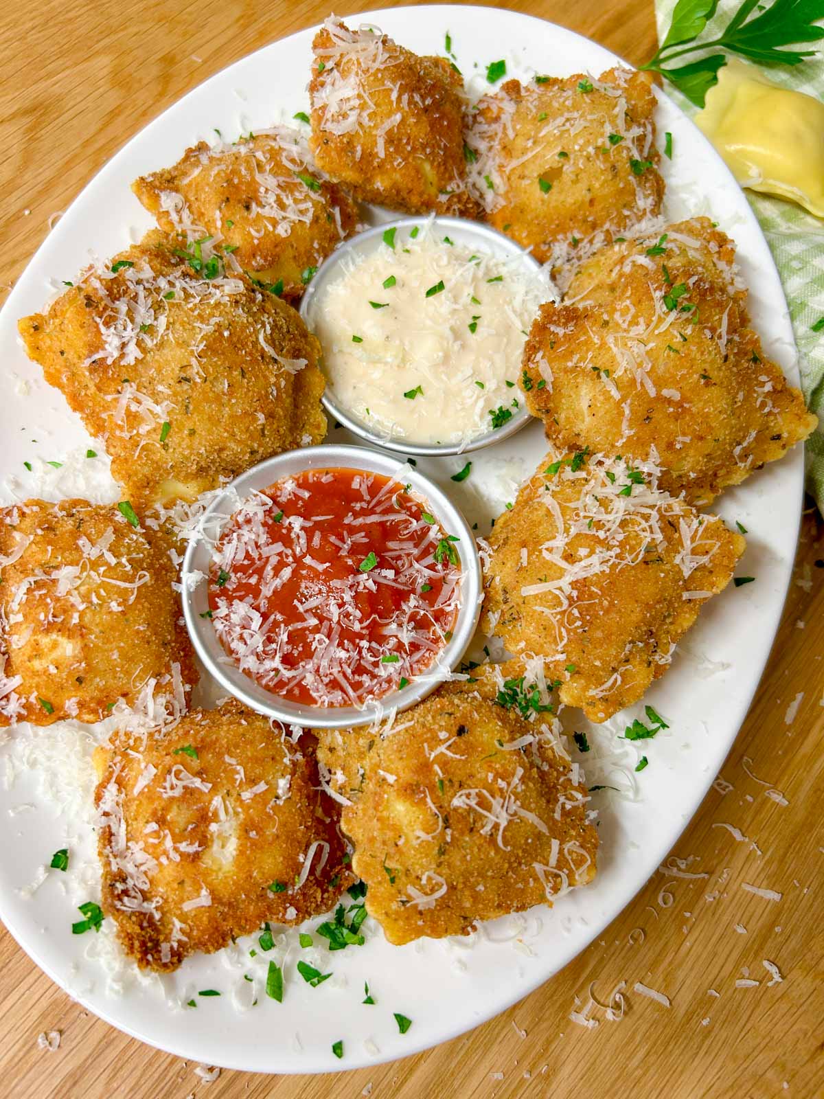 a white platter is filled with toasted ravioli, a bowl of marinara sauce, and a bowl of alfredo sauce. it is all topped with parmesan cheese and parsley