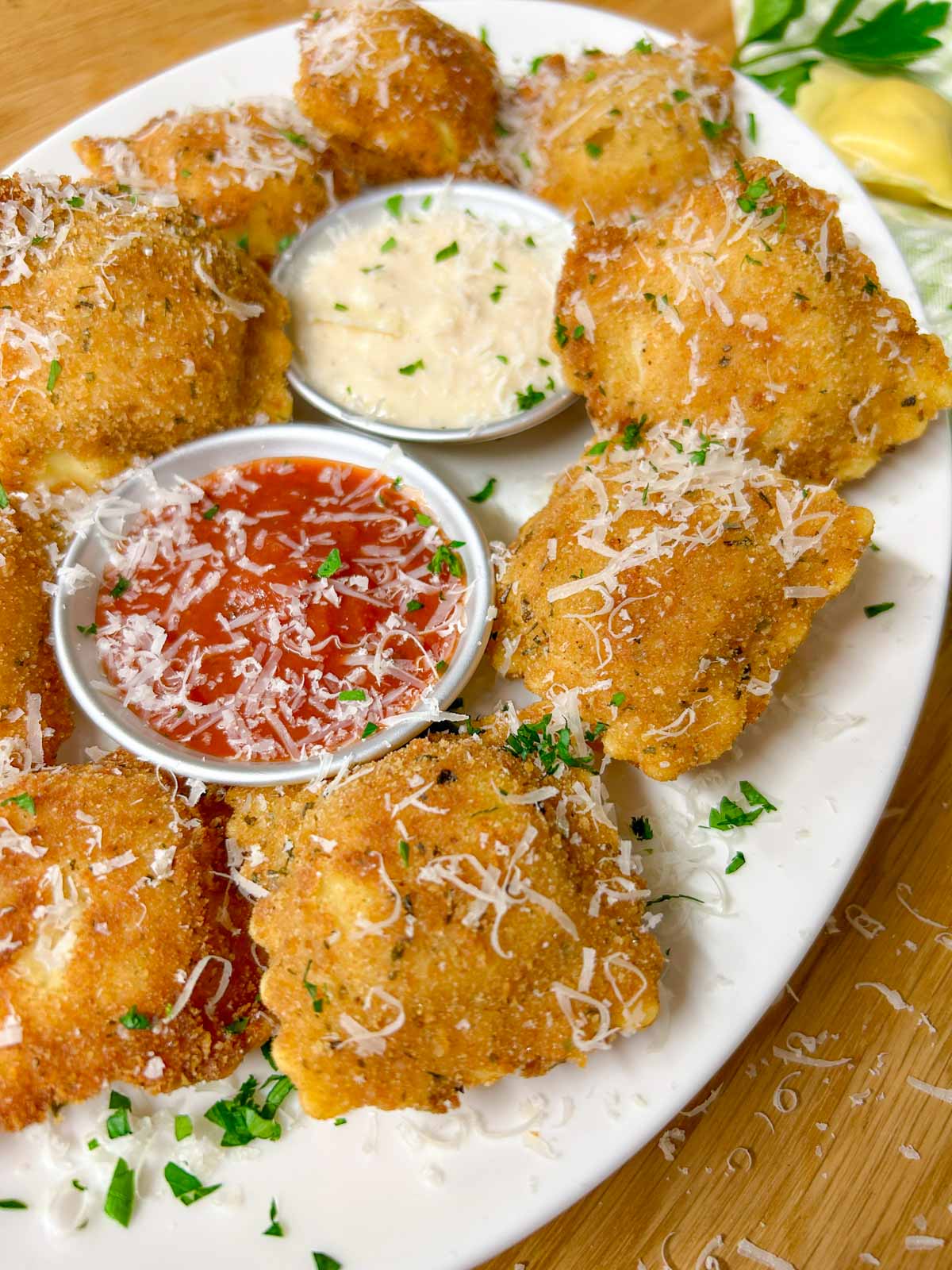 a white platter is filled with toasted ravioli, a bowl of marinara sauce, and a bowl of alfredo sauce. it is all topped with parmesan cheese and parsley