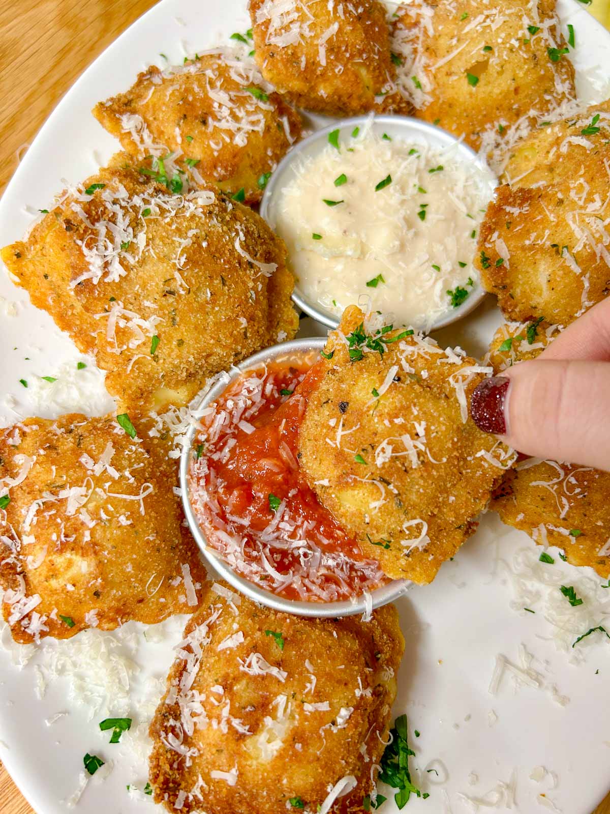 a white platter is filled with toasted ravioli, a bowl of marinara sauce, and a bowl of alfredo sauce. it is all topped with parmesan cheese and parsley. one is being dipped in the tomato sauce.