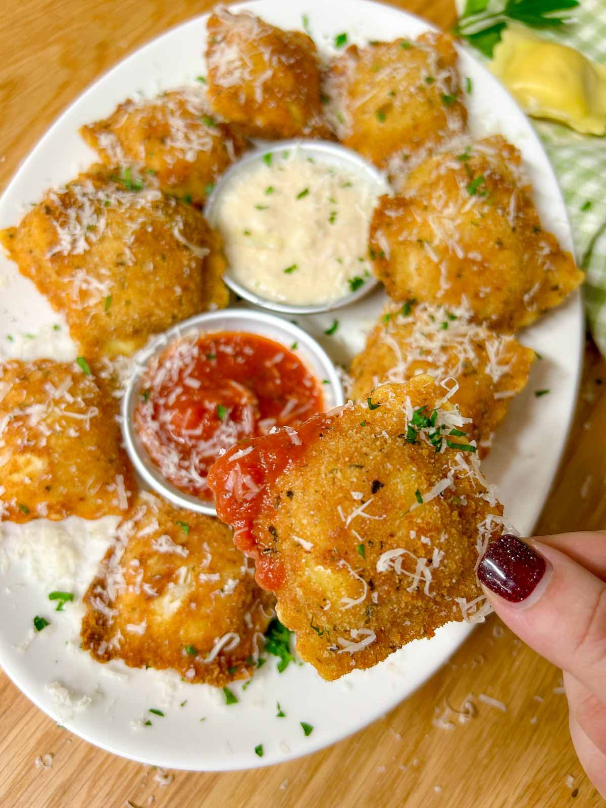 a white platter is filled with toasted ravioli, a bowl of marinara sauce, and a bowl of alfredo sauce. it is all topped with parmesan cheese and parsley. one is held up close to the camera