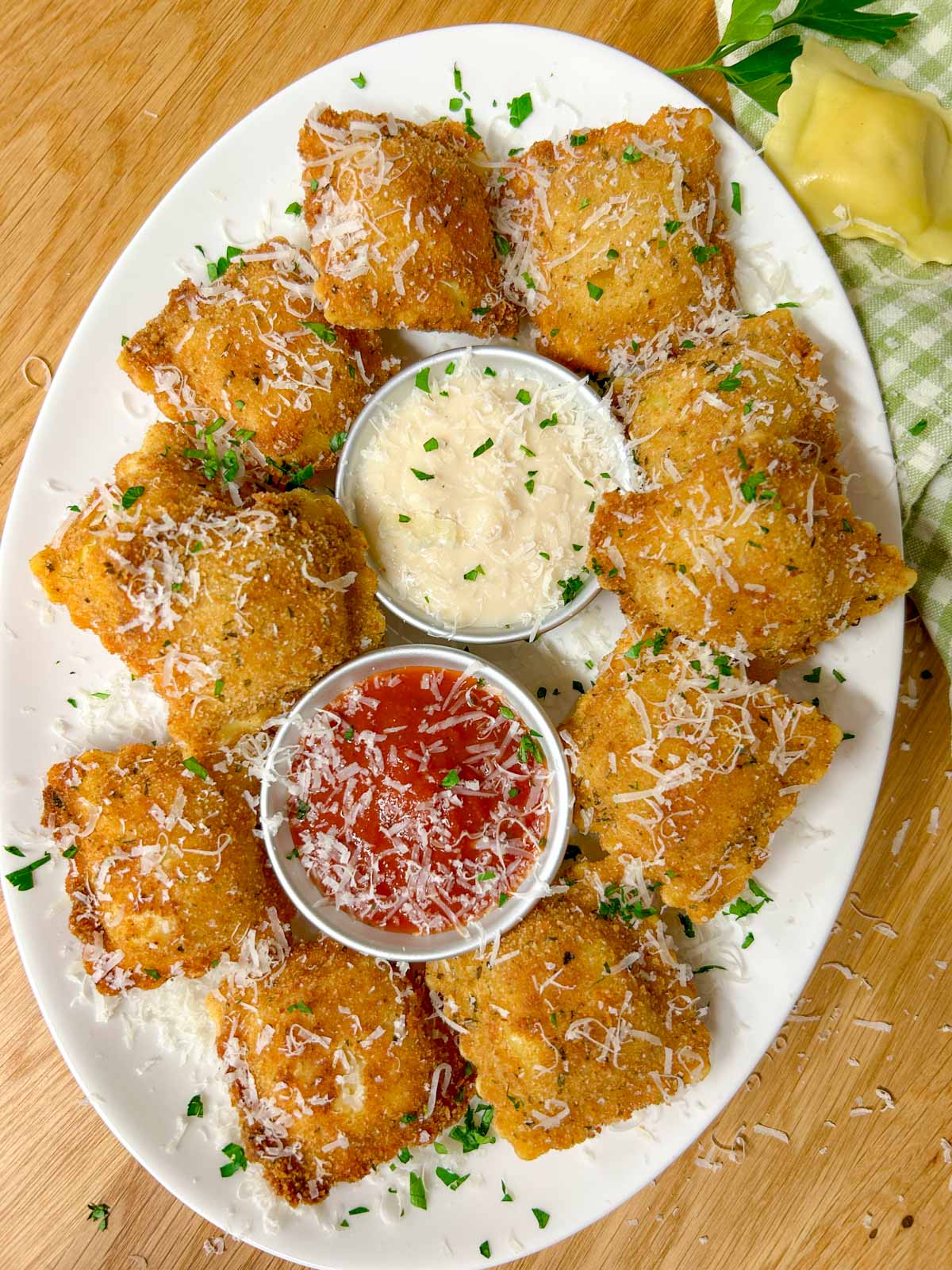 a white platter is filled with toasted ravioli, a bowl of marinara sauce, and a bowl of alfredo sauce. it is all topped with parmesan cheese and parsley