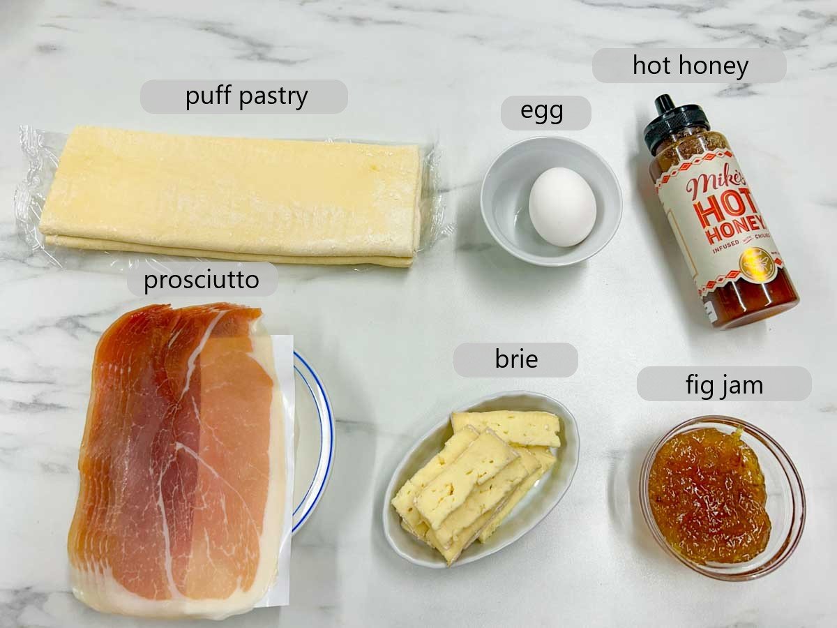 a kitchen counter with the ingredients to make prosciutto wrapped brie and fig puff with text overlays reading puff pastry, egg, hot honey, prosciutto, brie, and fig jam.
