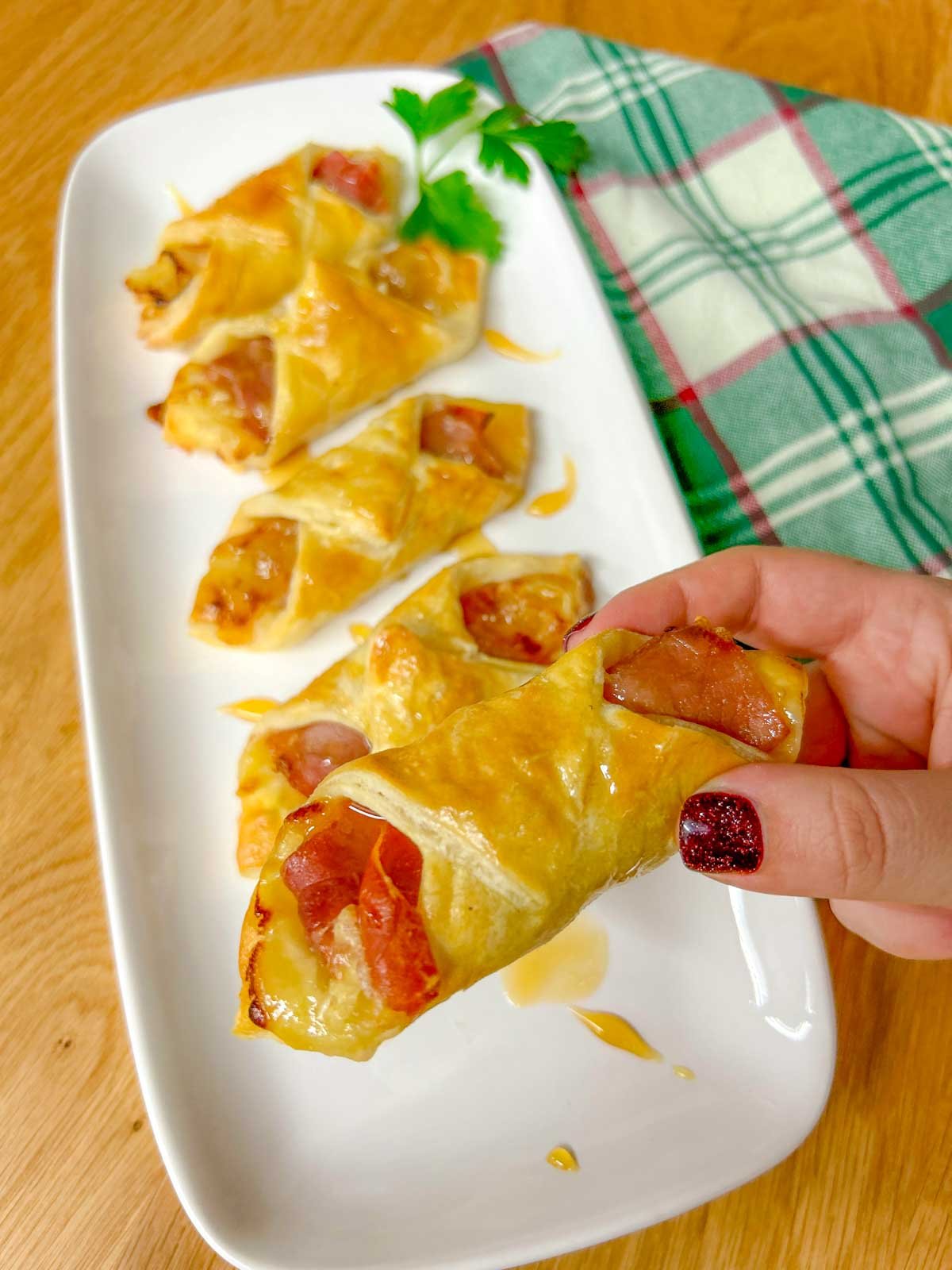 a white platter had prosciutto wrapped brie and fig puffs and they are drizzled with honey. one puff is held up close to the camera