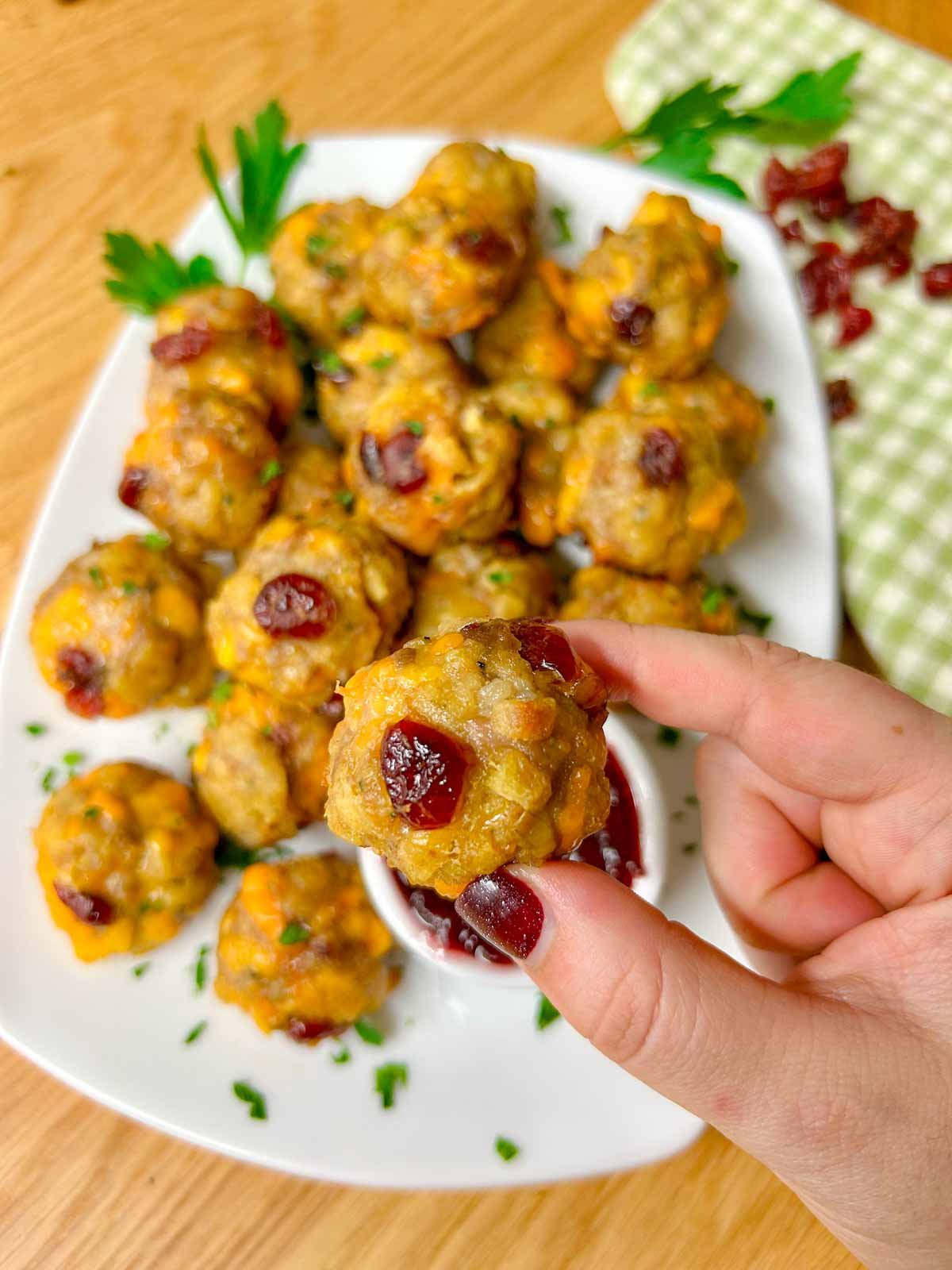 a white plate has sausage stuffing balls on top with a side dish of cranberry sauce for dipping. one ball is held up close to the camera