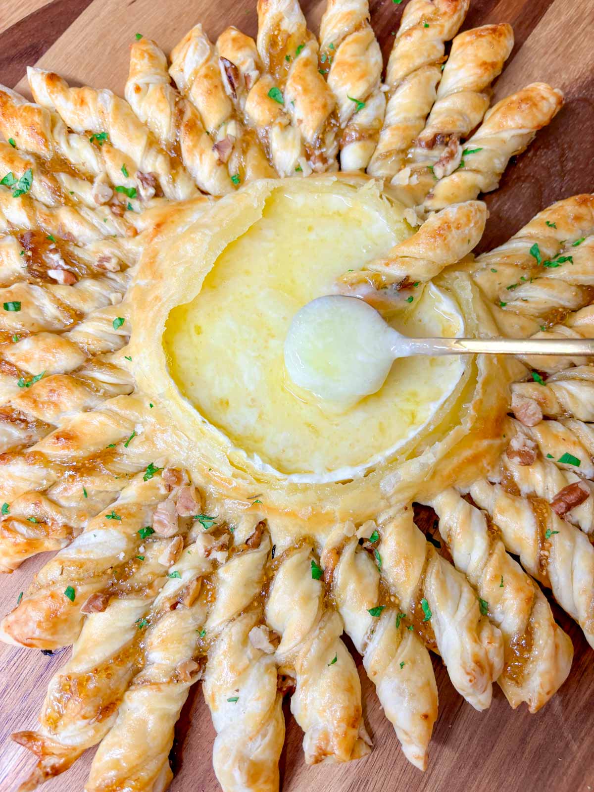 a wood serving board has a twisted pastry ring with a dipping bowl of brie in the centre. a spoon is dipped in the cheese