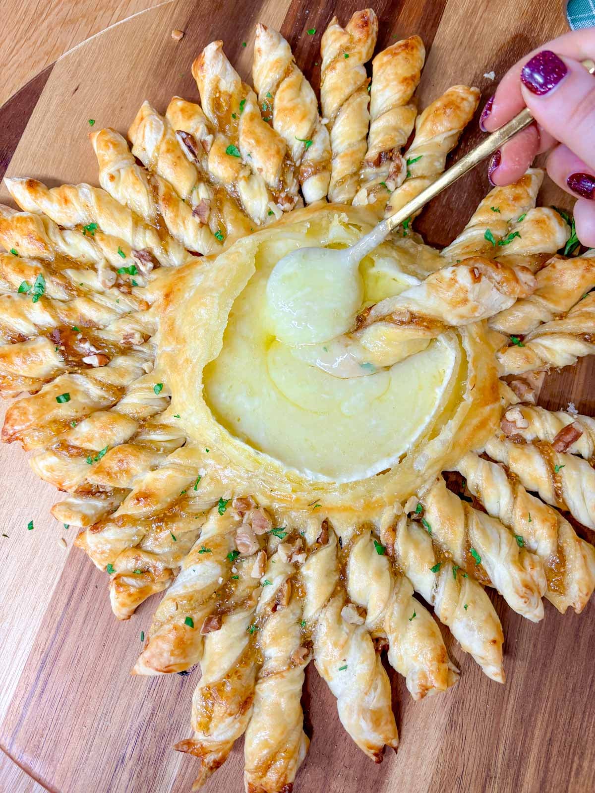 a wood serving board has a twisted pastry ring with a dipping bowl of brie in the centre. a spoon is dipped in the cheese