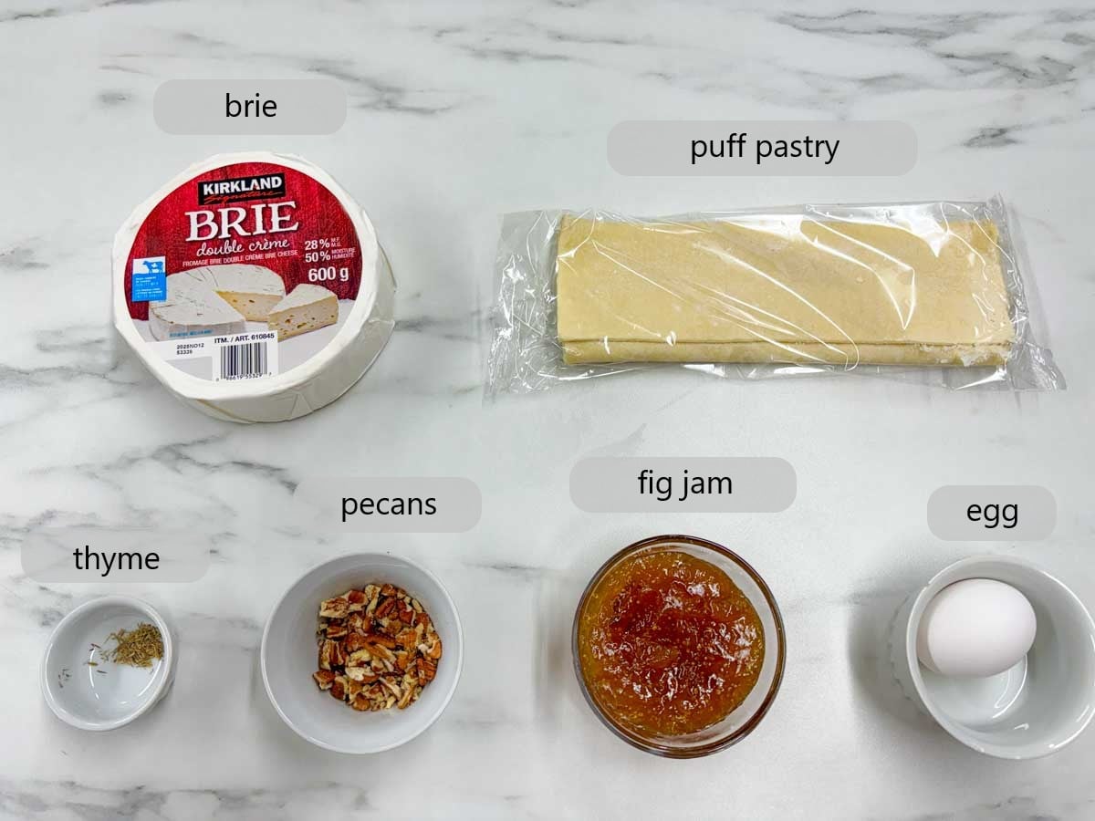 kitchen counter with bowls with ingredients and text overlays reading brie, puff pastry, thyme, pecans, fig jam and egg.