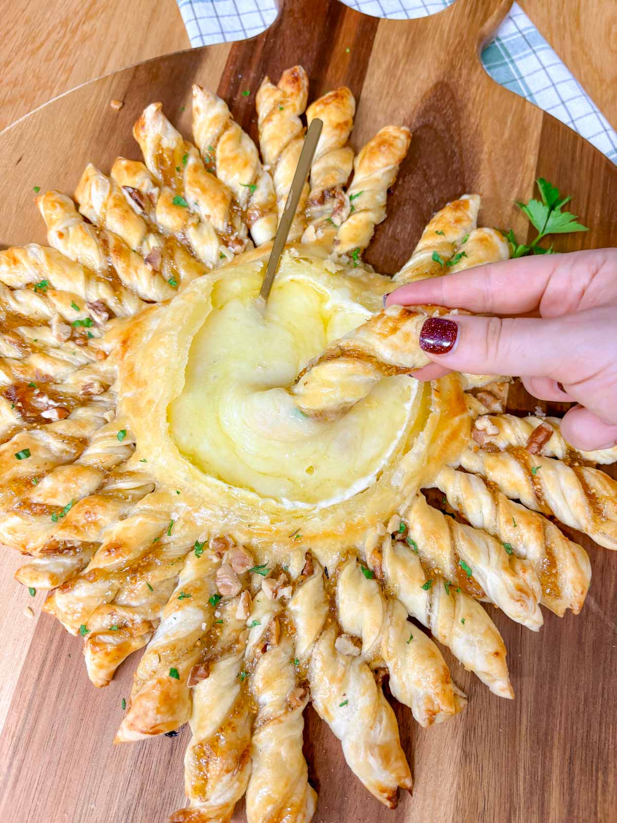 a wood serving board has a twisted pastry ring with a dipping bowl of brie in the centre. a twisted pastry is dipped in the cheese